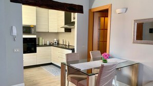 Una cocina moderna con armarios blancos, un microondas y un horno empotrado. Hay una mesa de comedor de vidrio con sillas, un jarrón de flores y un espejo en la pared.