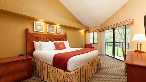 A hotel room with a large bed, a wooden nightstand, a desk, a chair, and a balcony with a view.