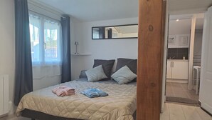 A bedroom with a bed, a bedside table, a mirror, and a view of the kitchen.