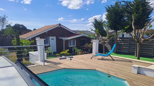 A swimming pool with a wooden deck, a blue lounge chair, and a house with a tiled roof.