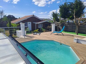 A swimming pool with a wooden deck, a blue lounge chair, and a house with a tiled roof.