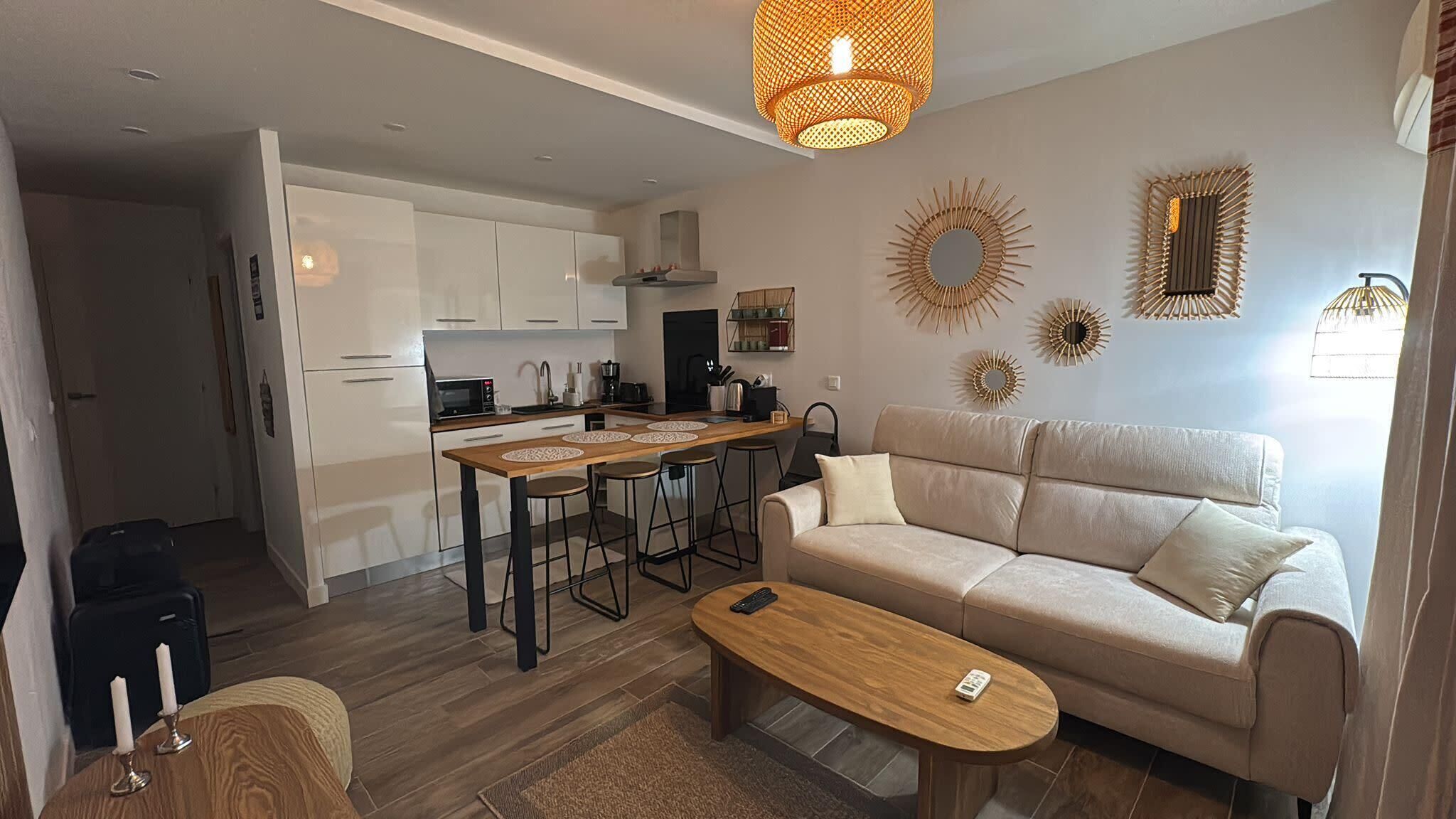 A modern living room with a sofa, a coffee table, a kitchen area with a dining table and bar stools, and decorative wall mirrors.