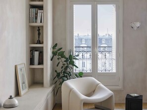 A room with a white chair, a bookshelf, a potted plant, and a view of buildings through a window. - Spacious apartment overlooking the Eiffel Tower (Paris)