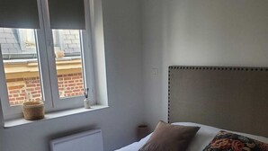 Ein Schlafzimmer mit Bett, Fenster mit Jalousien, Heizkörper und einem an der Wand befestigten Kopfteil.