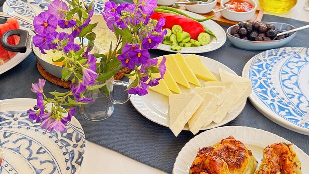 Mesa posta com diversos pratos, incluindo pão, queijo e uma salada, em uma sala de jantar com vista para o verde.