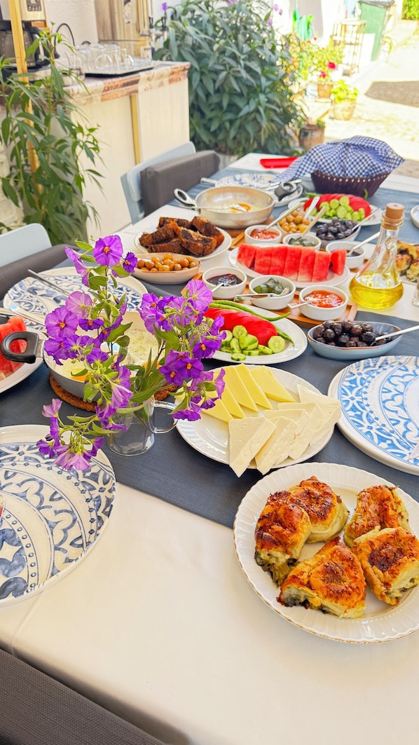 A table set with various dishes, including bread, cheese, and a salad, in a dining area with a view of greenery. - Şenol Pansiyon (Bozcaada)