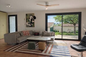 Un salon comprenant un canapé d’angle, une table basse ronde, un fauteuil en cuir noir et une grande porte-fenêtre coulissante donnant sur un jardin.