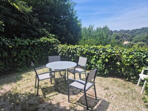 Un espace extérieur aménagé avec une table ronde et quatre chaises, entouré d’une végétation luxuriante et d’une haie.
