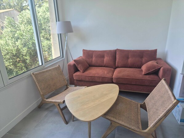 Une pièce avec un canapé rouge, deux fauteuils en osier, une petite table ronde, un lampadaire et une grande fenêtre.