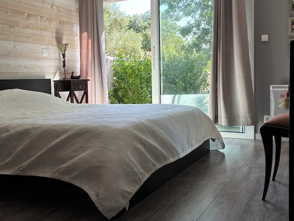 A bedroom with a large bed, a wooden headboard, a nightstand, a lamp, and a view of greenery outside. - House with private pool and large garden (Nieul-sur-Mer)