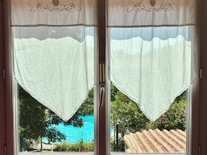 A window with a white curtain and a view of a blue pool and greenery. - House with private pool and large garden (Nieul-sur-Mer)