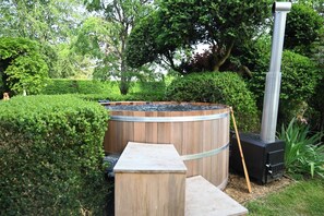 A hot tub surrounded by a wooden barrel and a chimney, set in a garden with hedges and trees. - Luxury home in the Country with Cedar Hot Tub, Tennis Court and Gym (Midgham)