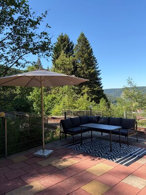 An outdoor patio with a table and a sofa, a large umbrella, and a patterned rug.