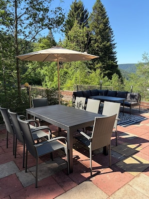An outdoor patio with a table and chairs, a large umbrella, and a view of trees and hills.