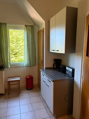 A compact kitchen with a window, a small stool, a red trash bin, and a coffee maker.