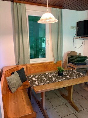 A wooden bench with cushions, a table with a floral tablecloth, a hanging light fixture, a television on a wall, and a window with shutters.