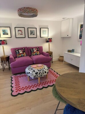 A living room with a purple sofa, a round wooden table, a patterned ottoman, and a wall with framed artwork.