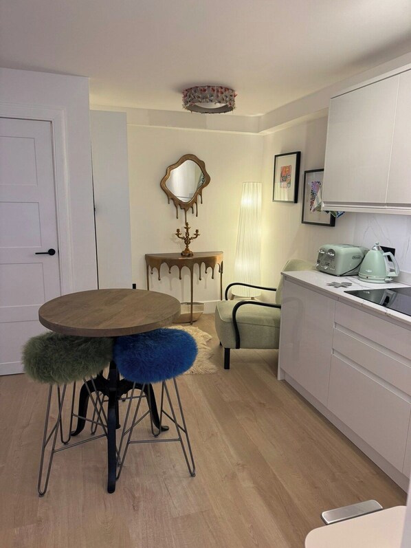 A compact kitchen with a round wooden table, a chair with a fluffy seat, a small console table with a mirror, and a white cabinet with a toaster and kettle.