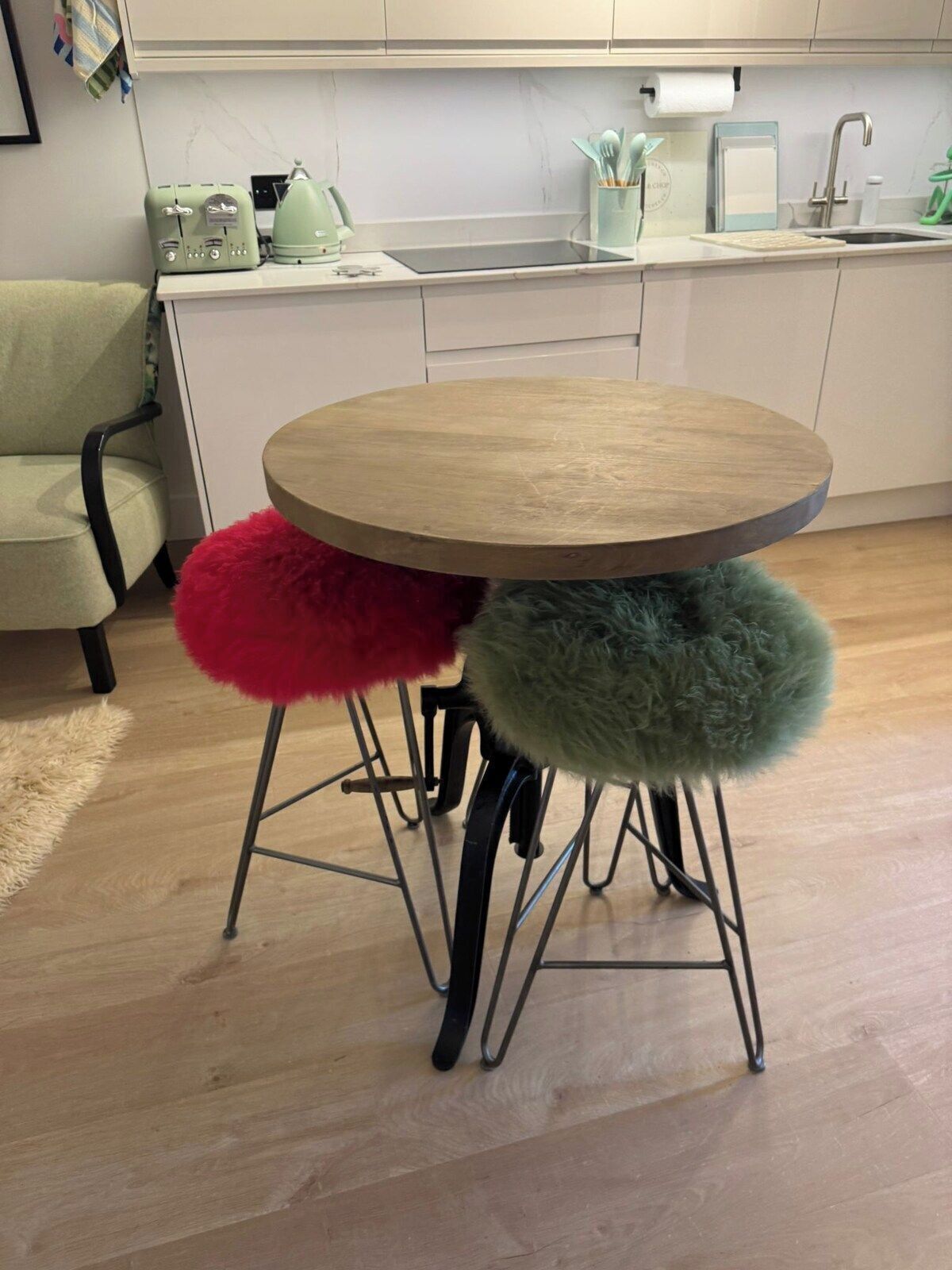 A round wooden table with metal legs, two stools with fluffy cushions, a kitchen area with a sink, and a chair.