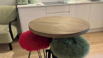 A round wooden table with metal legs, two stools with fluffy cushions, a kitchen area with a sink, and a chair.