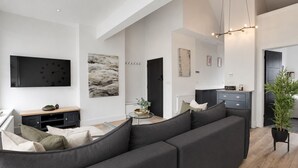 A modern living room with a sectional sofa, a flat-screen TV, abstract art, and a dining area with a kitchen in the background. - Stylish 1BR Penthouse with Balcony - City Centre near Cliffs Pavillion (Southend-on-Sea)