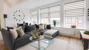 A modern living room with a large wall clock, a sofa with cushions, a glass coffee table, and large windows with blinds. - Stylish 1BR Penthouse with Balcony - City Centre near Cliffs Pavillion (Southend-on-Sea)