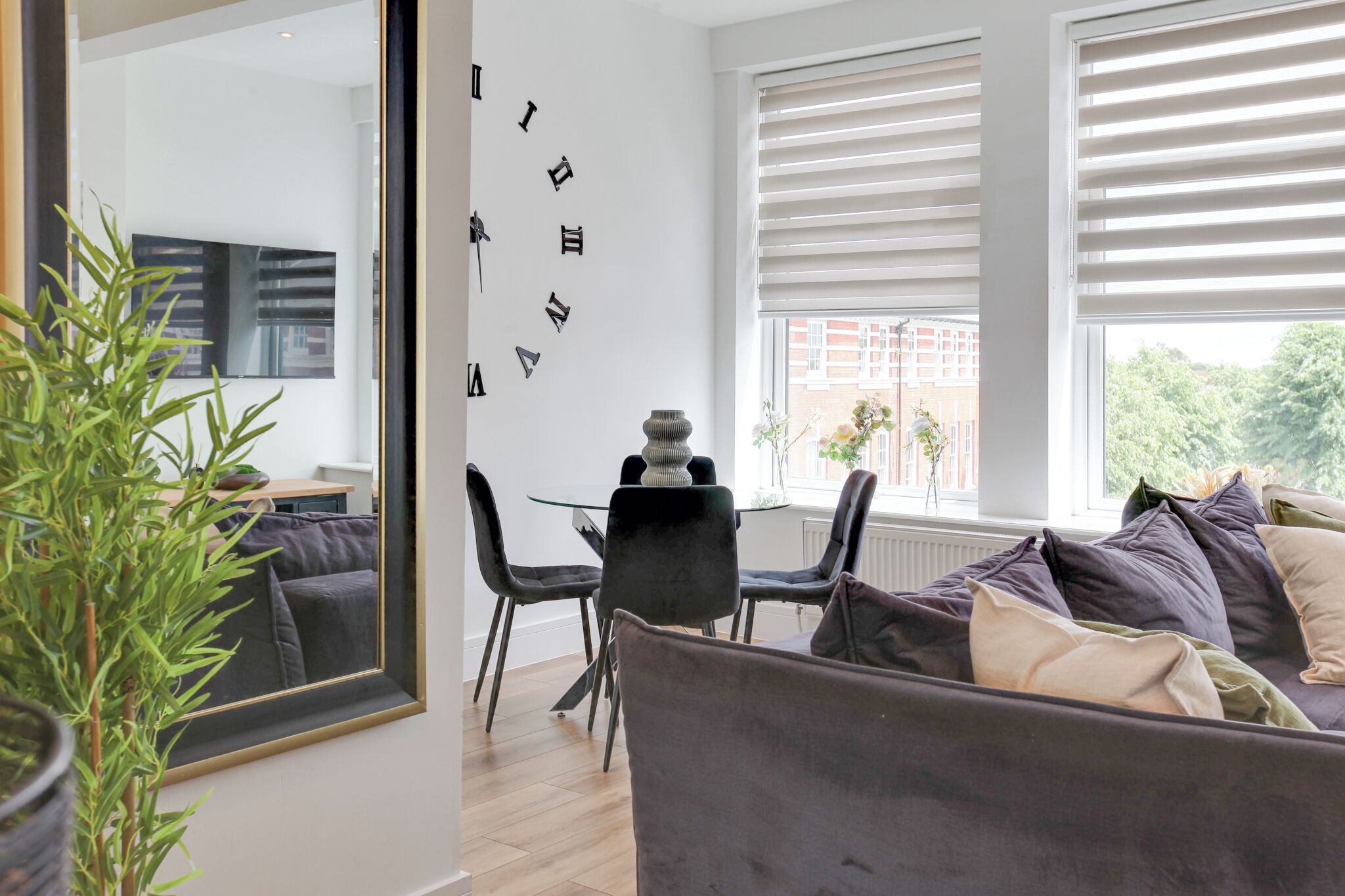 A modern living room with a sofa, a dining table, chairs, a large mirror, and a window with blinds.