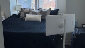 Un dormitorio con cama, almohadas, un escritorio, un ventilador y vistas de la ciudad a través de amplias ventanas.