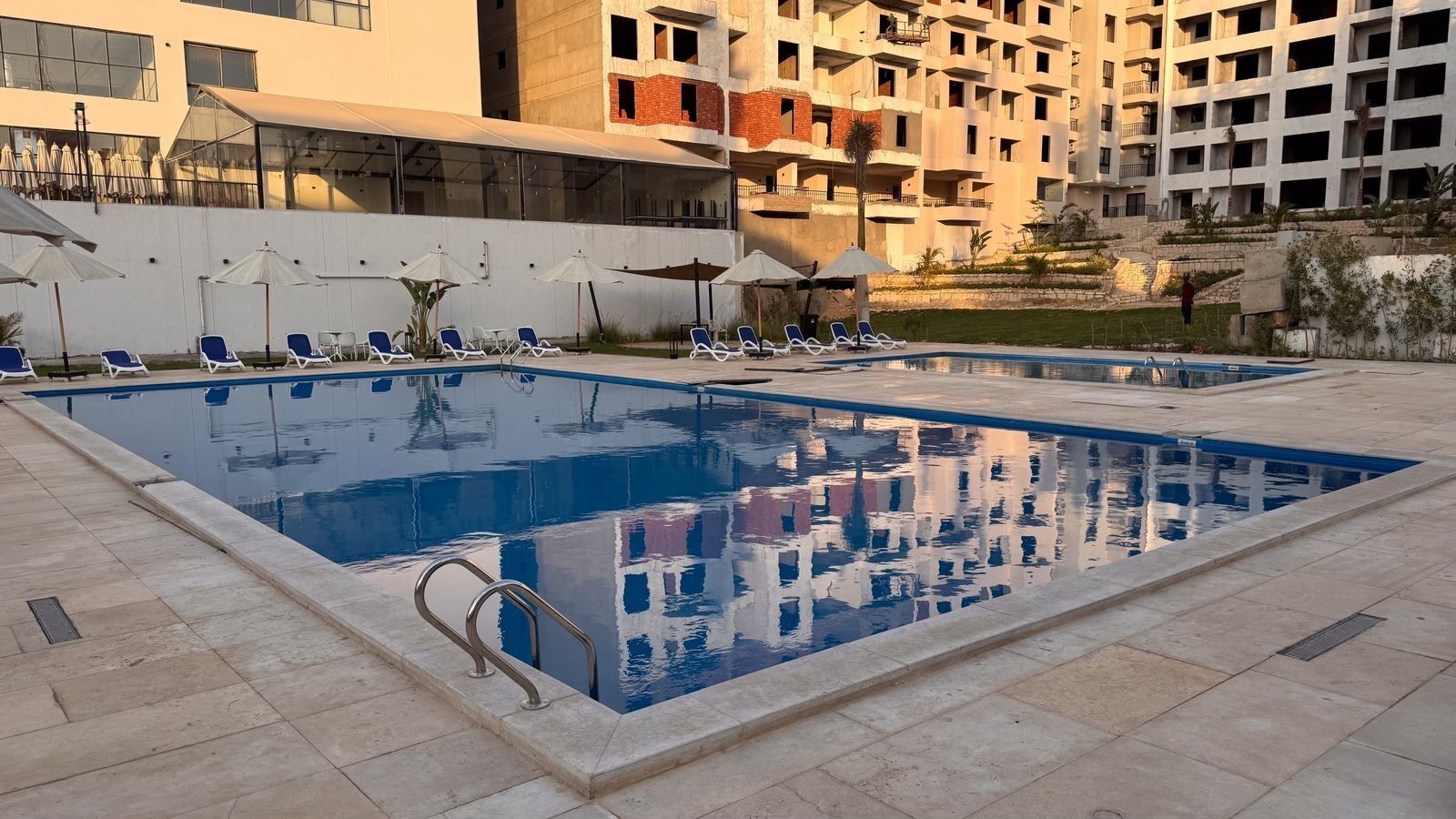 Piscina con sillas de descanso y sombrillas, rodeada por un edificio con balcones.