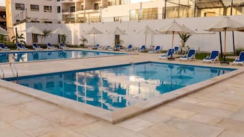 Piscina con sillones y sombrillas, rodeada por un edificio con balcones.