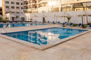 Piscina con sillones y sombrillas, rodeada por un edificio con balcones.
