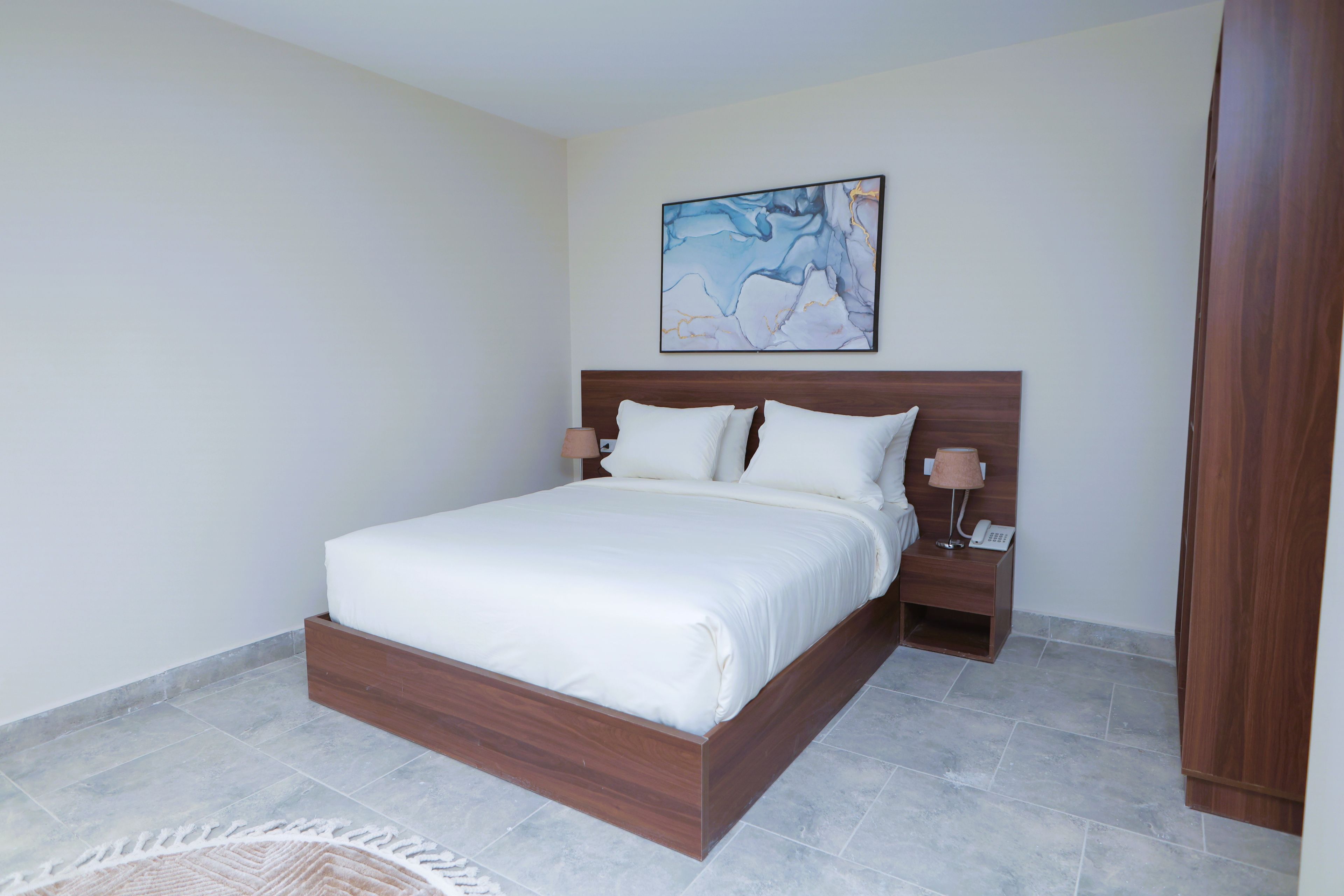 Un dormitorio con estructura de cama de madera, ropa de cama blanca, una mesita de noche con lámpara y un cuadro en la pared.