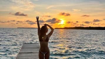 A person standing on a wooden pier extending into a body of water, with a sunset in the background.