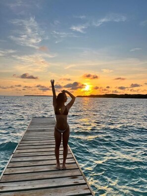 A person standing on a wooden pier extending into a body of water, with a sunset in the background. - Orika del Mar  (Rosario Islands)