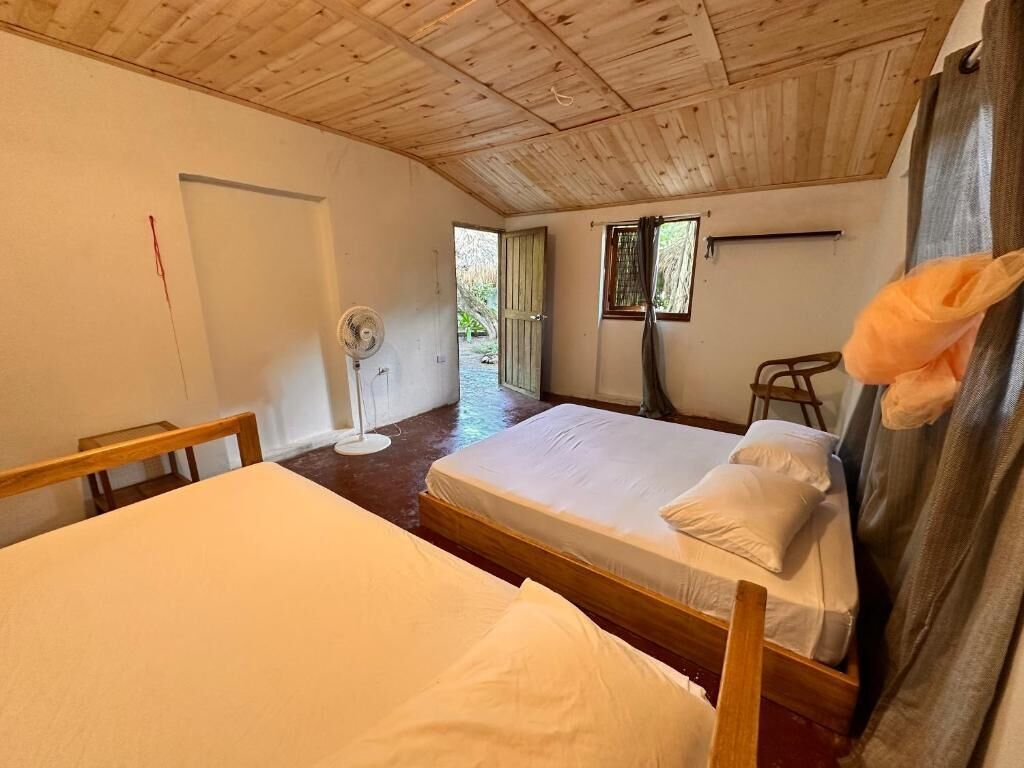 Quarto com duas camas, um ventilador, uma janela e uma porta.