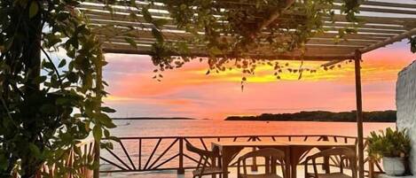 An outdoor seating area with a pergola, a table, and chairs, overlooking a sunset over a body of water.