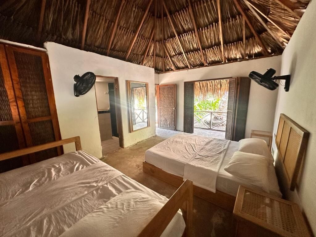 A room with two beds, a thatched roof, and a view of greenery outside.