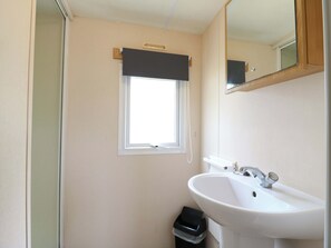 A bathroom with a white sink, a mirror, a window with a black blind, and a small trash bin. - Caravan 102 Seawick (Clacton-on-Sea)