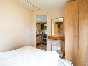 A bedroom with a bed, a wooden wardrobe, a small stool, and a kitchenette in the background. - Caravan 102 Seawick (Clacton-on-Sea)