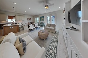 A spacious living area with a sofa, ottoman, and chairs, a kitchen with white cabinets, and a large TV mounted on the wall. - Tiki Pool Club (Galveston)