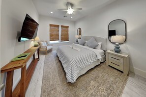 Une chambre à coucher comprenant un lit, des tables de chevet, un téléviseur, un ventilateur de plafond et une fenêtre avec des stores.
