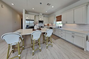 A modern kitchen with white cabinets, a central island with bar stools, and stainless steel appliances. - Tiki Pool Club (Galveston)