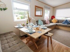A cozy dining area with a wooden table set for two, a sofa with cushions, and a window with blinds. - Foxglove 4 (Ringwood)
