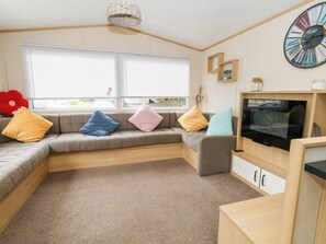 A modern living room with a sectional sofa, a flat-screen TV, a large wall clock, and a window with blinds. - Foxglove 4 (Ringwood)