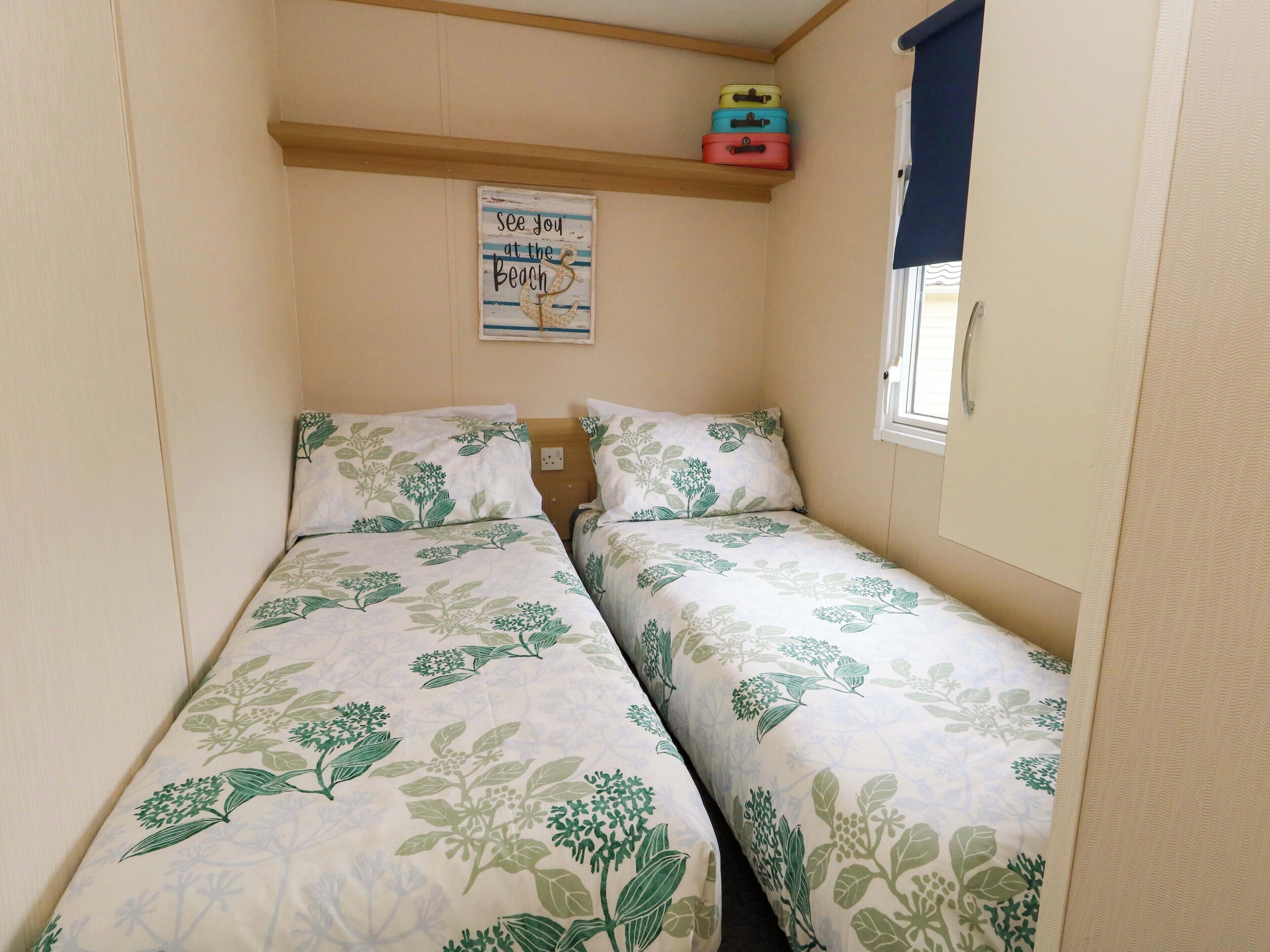 A small, cozy room with two beds, a wall-mounted shelf with colorful suitcases, a window with a curtain, and a sign on the wall.