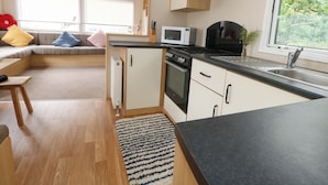 A compact kitchen with a microwave, stove, and sink, a small dining area with a table and chairs, and a window with a view of greenery. - Foxglove 4 (Ringwood)