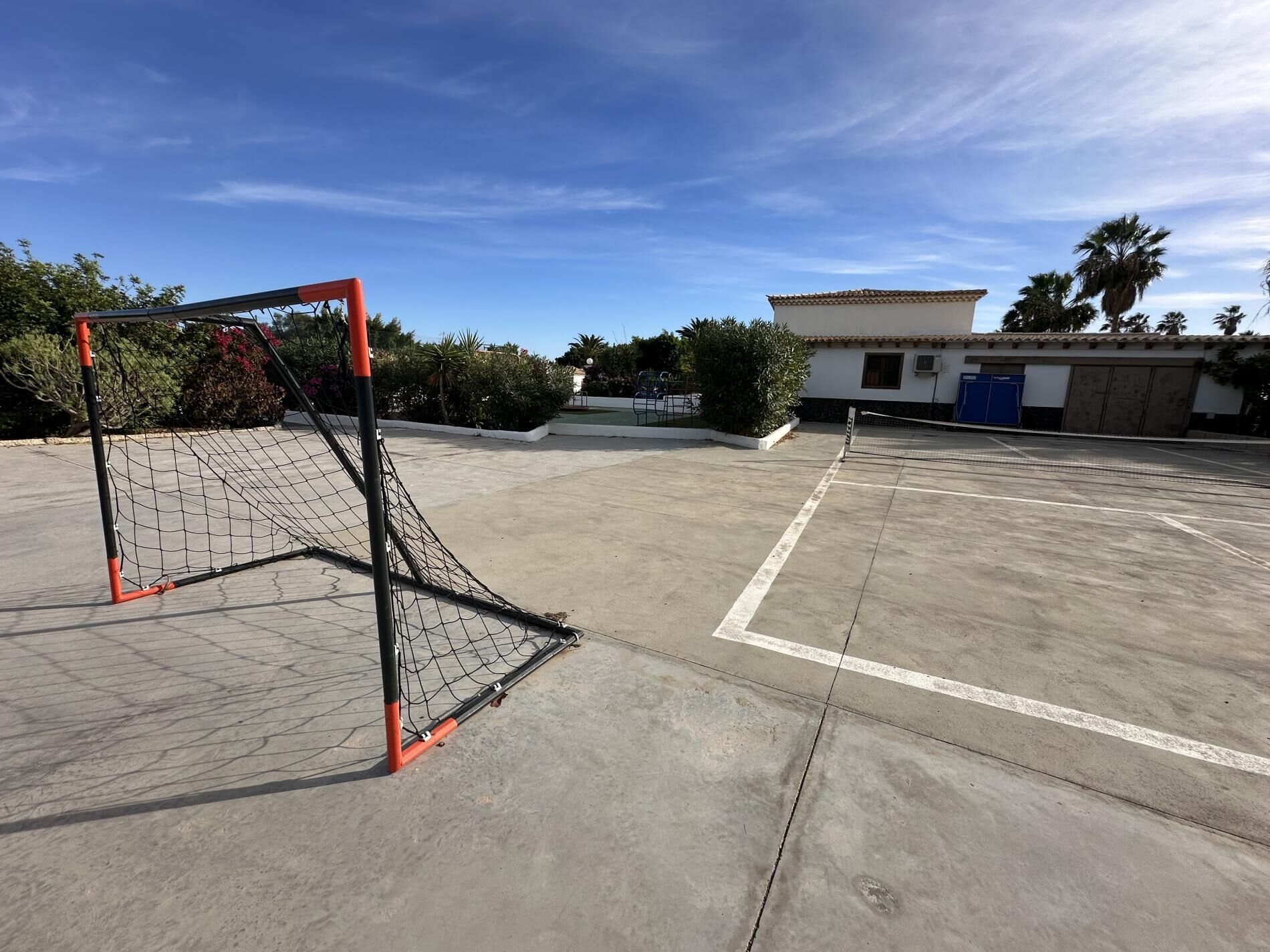 Un terrain de sport extérieur avec des poteaux de but, un bâtiment doté d’un toit en tuiles et une végétation luxuriante tout autour.