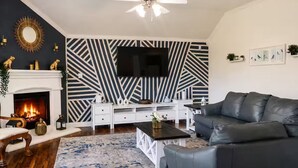 A living room with a fireplace, a black and white patterned wall, a flat-screen TV, a black leather sofa, and a coffee table. - Executive 4BR Retreat Mins to AT&T Stadium (Arlington)