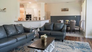 A living room with two grey sofas, a coffee table, and a dining area with a table and chairs. - Executive 4BR Retreat Mins to AT&T Stadium (Arlington)