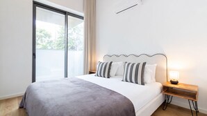 Une chambre d’hôtel moderne avec un grand lit, une table de chevet en bois, une lampe et une porte-fenêtre coulissante donnant sur un espace extérieur.
